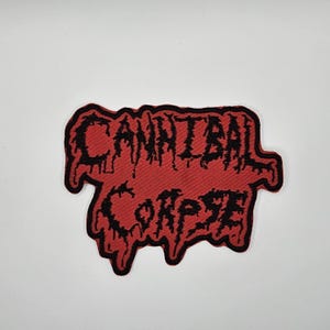 May include: A red patch featuring the band name "CANNIBAL CORPSE" in black, dripping-style lettering. The patch has a black border. This patch is designed to be sewn or ironed onto clothing or accessories, and has a distressed aesthetic.