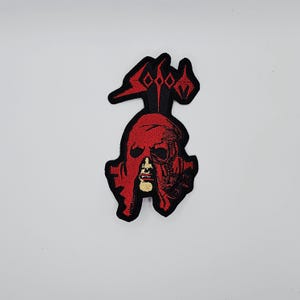 Könnte beinhalten: Gestickter Aufnäher mit einem roten, stilisierten Schädeldesign und schwarzen Akzenten. Der Aufnäher hat ein detailliertes, strukturiertes Aussehen mit einem goldfarbenen Element in der Mitte. Das Design ist vor einem weißen Hintergrund platziert.