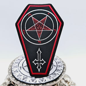 Può includere: Una toppa ricamata nera e rossa a forma di bara con un pentagramma e una testa di capra all'interno. Il pentagramma è circondato da un cerchio con caratteri ebraici.