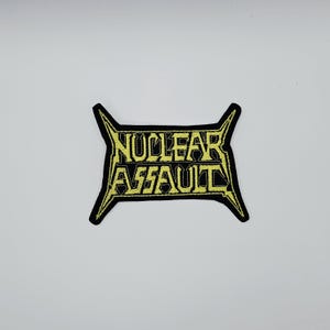 Peut inclure: Un écusson brodé noir avec les mots "NUCLEAR ASSAULT" en jaune, avec une typographie stylisée. L'écusson a une forme unique avec des bords pointus s'étendant sur les côtés. Il est probablement destiné à une veste ou un sac.