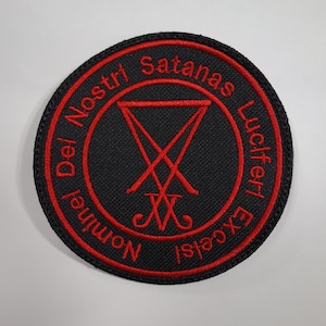 Puede incluir: Un parche redondo negro con texto bordado en rojo y un símbolo rojo. El texto rodea el símbolo y dice "Dei Nostri Satanas Luciferi Excelsi Nominel". El símbolo central es una V invertida estilizada con una doble cruz en la parte inferior.
