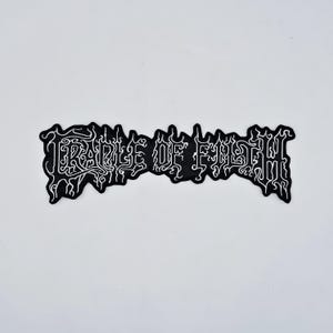 Op de afbeelding: Een zwart-witte patch met de tekst "CRADLE OF FILTH" in een gestileerd, gotisch lettertype. De witte tekst staat op een zwarte achtergrond. De patch heeft een unieke, onregelmatige vorm.
