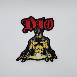 Puede incluir: Parche bordado con la palabra "DIO" en letras góticas rojas. Debajo, una figura negra estilizada con detalles amarillos y dorados, y cadenas. Ideal para personalizar ropa o bolsos.