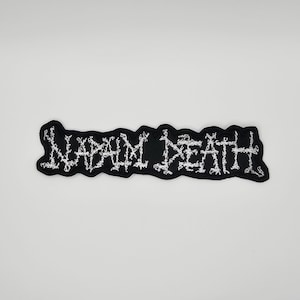 May include: A black patch with the white text "NAPALM DEATH" in a stylized, jagged font. The patch has a black border and is likely made of fabric, suitable for sewing or ironing onto clothing or accessories.