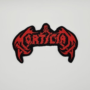 Puede incluir: Un parche bordado en negro y rojo que presenta la palabra MORTICIAN en una fuente estilizada y dentada. El diseño incluye un pequeño icono de calavera centrado sobre el texto, con extensiones afiladas en forma de ala a ambos lados.