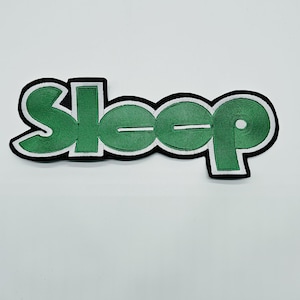 May include: An embroidered patch featuring the word Sleep in green lettering with a white outline and a thick black border. The text is stylized in a bold, rounded font, creating a cohesive graphic design suitable for application onto fabric items.