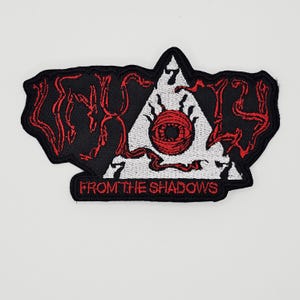 May include: Black patch with red lettering and a white triangle featuring a red eye design. The patch reads "FROM THE SHADOWS" at the bottom. The patch has a unique, irregular shape.