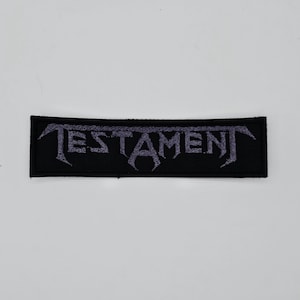 Op de afbeelding: Zwart rechthoekig embleem met het woord "TESTAMENT" in paars geborduurd. De tekst is in een gotisch lettertype. Het embleem is waarschijnlijk voor een jas of tas en is een bandmerchandise-artikel.