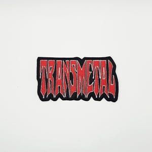 May include: Embroidered patch with the word "TRANSMETAL" in red, outlined in white, on a black background. The text has a bold, stylized font. The patch is rectangular with a wavy border.