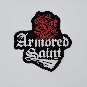 以下が含まれることがあります： 白いゴシック体の文字で「Armored Saint」と書かれた黒い刺繍パッチ。パッチには、テキストの上に赤いヘルメットをかぶった顔のイラストが描かれています。パッチには黒い縁取りがあります。