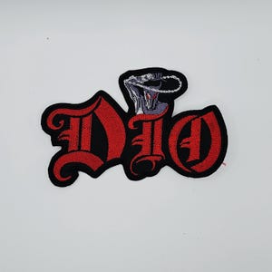 May include: Embroidered patch featuring the word "DIO" in a gothic font, with red letters outlined in black. Above the letters is a gray and white illustration of a figure with a chain. The patch is likely for clothing or accessories.