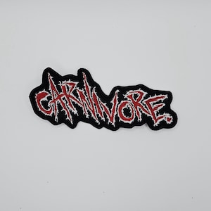 May include: A black embroidered patch with the word "CARNIVORE" in red, stylized, jagged lettering. The patch has a black border and a white outline around the red text, creating a bold contrast.