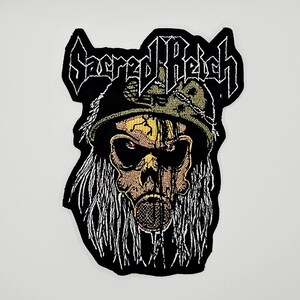 May include: An embroidered patch featuring a skull wearing a military-style helmet and a gas mask, with long white hair. The text Sacred Reich is stitched in a bold, stylized font at the top. The design uses black, white, tan, and yellow thread.