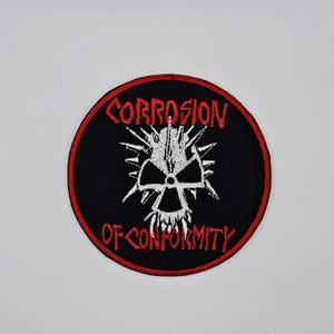 Peut inclure: Écusson rond noir avec une bordure rouge. Le patch présente le texte "CORROSION OF CONFORMITY" en rouge, entourant un motif de crâne blanc avec des pointes.