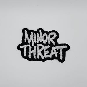 Könnte beinhalten: Schwarz-weißer Aufnäher mit dem Schriftzug "MINOR THREAT" in weißer, stilisierter Schrift. Der Aufnäher hat einen schwarzen Rand und ist zum Aufnähen oder Aufbügeln auf Kleidung oder Accessoires gedacht.