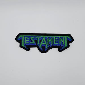 May include: Embroidered patch with the word "TESTAMENT" in stylized green and blue lettering. The patch has a black border and a unique, jagged shape. The design is set against a white background.