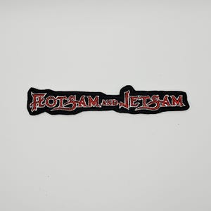 Puede incluir: Un parche de tela bordado que presenta el texto Flotsam and Jetsam en una fuente roja estilizada con un contorno negro. Las letras tienen un diseño afilado y angular, y todo el parche está bordeado por un grueso borde negro sobre un fondo liso.