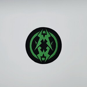 Puede incluir: Un parche redondo negro con un emblema verde estilizado. El emblema presenta un diseño geométrico con formas de rayo y una figura central en forma de cruz. El parche es probablemente un accesorio para planchar o coser.
