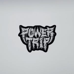 May include: A black and white embroidered patch featuring the text "POWER TRIP" in a stylized, gothic font. The patch has a jagged, irregular border and is set against a white background.