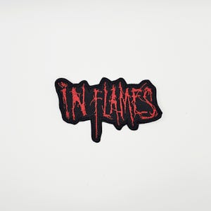 Op de afbeelding: Een zwart embleem met de rode geborduurde tekst "IN FLAMES". De letters zijn in een gekarteld, gestileerd lettertype. De patch heeft een zwarte rand en wordt weergegeven op een witte achtergrond.