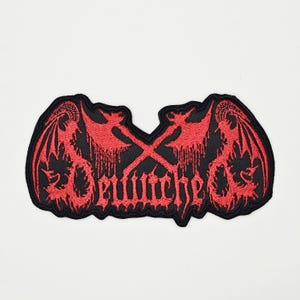 Könnte beinhalten: Schwarzer Aufnäher mit rotem Stickdesign. Das Design zeigt gekreuzte Äxte, stilisierte Flügel und das Wort "Bewitcher" in einer gotischen Schriftart. Der Aufnäher hat einen schwarzen Rand und ist wahrscheinlich für Kleidung oder Accessoires.