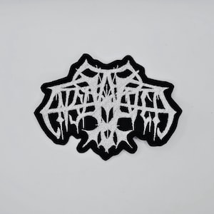 May include: Embroidered patch with a black border and white, stylized text. The text appears to be a band logo or similar design, with a gothic or metal aesthetic. The patch has a jagged, irregular shape.