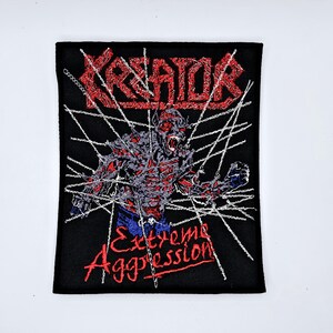 以下が含まれることがあります： 赤色の「KREATOR」の文字が刺繍されたパッチ。パッチには、赤色の「Extreme Aggression」という文字とモンスターのグラフィックが描かれています。背景は黒で、銀色の鎖のような線があります。