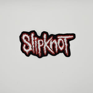 Puede incluir: Un parche de tela bordado que presenta la palabra Slipknot en una fuente blanca, estilizada y dentada. Las letras están delineadas con hilo rojo y colocadas sobre un fondo negro sólido, creando una apariencia texturizada de alto contraste.
