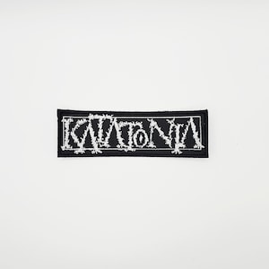 Könnte beinhalten: Ein schwarzer, rechteckiger Aufnäher mit dem Wort "KATATONIA" in weißer, stilisierter gotischer Schrift gestickt. Der Aufnäher, für Kleidung oder Accessoires, vor weißem Hintergrund.