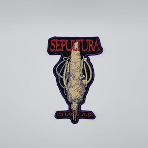 以下が含まれることがあります： 黒と紫の背景に赤い文字でバンド名「SEPULTURA」が刺繍されたパッチ。 パッチの中央にはチューブの付いた包まれた人物のグラフィックがあり、その下に「CHAOS A.D.」の文字があります。