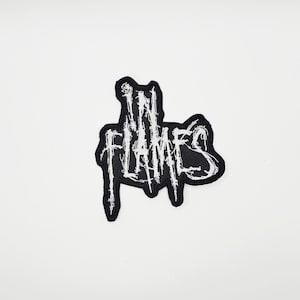 May include: Black and white embroidered patch with the words "IN FLAMES" in a stylized, distressed font. The patch has a jagged, irregular border and is likely designed to be sewn or ironed onto fabric.