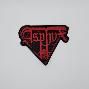 Puede incluir: Un parche negro con la palabra "Asphyx" en letras góticas rojas. El parche tiene forma triangular con un gráfico de calavera en la parte inferior. Probablemente es para una chaqueta o bolso.