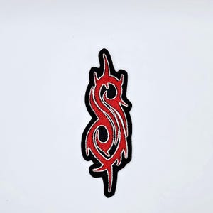 Puede incluir: Parche bordado con un diseño rojo y detalles blancos sobre fondo negro. El diseño es una forma vertical en forma de llama con curvas intrincadas y bordes puntiagudos. Para ropa o accesorios.