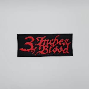 May include: Black rectangular patch with the words "3 Inches of Blood" embroidered in red. The text is in a gothic, stylized font. The patch is likely for a jacket or bag.