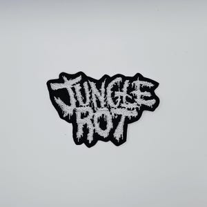 May include: A black and white embroidered patch with the text "JUNGLE ROT" in a distressed, dripping font. The patch has a black border and is set against a white background.