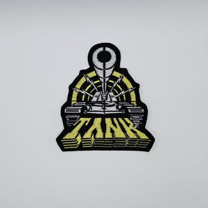Könnte beinhalten: Gestickter Aufnäher mit einem stilisierten Panzer-Design in Schwarz, Gelb und Weiß. Das Wort "TANK" ist in einer kräftigen, gelben Schrift am unteren Rand zu sehen. Der Aufnäher hat einen schwarzen Rand.