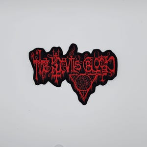 May include: A black patch with red embroidered text that reads "The Devil's Blood". The patch features a stylized design with a pentagram and other symbols. The patch is likely for clothing or accessories.