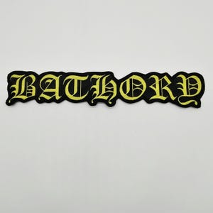 May include: An embroidered patch featuring the word BATHORY in a yellow gothic-style font. The lettering is stitched onto a black fabric base that follows the contour of the letters, creating a distinct, bold, and high-contrast visual design.