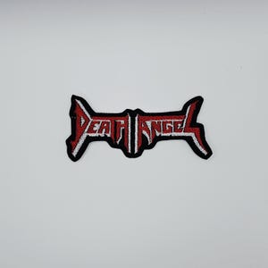 Könnte beinhalten: Gestickter Aufnäher mit dem Text "DEATH ANGEL" in einer stilisierten, gotischen Schriftart. Der Text ist rot mit weißen Umrissen, vor einem schwarzen Hintergrund. Der Aufnäher hat eine einzigartige, eckige Form.