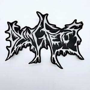 May include: A black and white embroidered patch with a stylized design. The patch features a black background with a white, intricate design that appears to be a word or symbol. The edges are jagged and irregular.