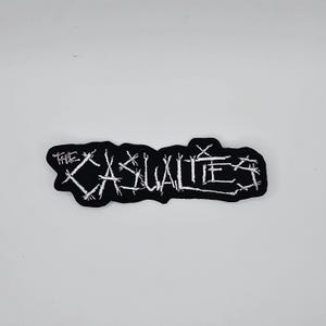 Puede incluir: Parche bordado negro con el texto blanco "THE CASUALTIES". El parche tiene un borde negro y está diseñado para ser cosido o planchado en ropa o accesorios.