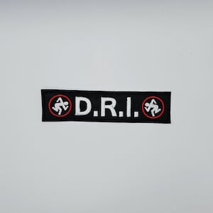 Op de afbeelding: Zwart rechthoekig embleem met de witte letters "D.R.I.". Elk uiteinde van het embleem heeft een rood en wit cirkelvormig embleem met een stokfiguur. Dit embleem is waarschijnlijk een accessoire voor kleding of tassen.