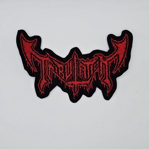 May include: A black embroidered patch with the word "TRIBULATION" in red, gothic-style lettering. The patch has a unique, stylized shape with pointed edges and a dripping effect, suitable for jackets, bags, or other apparel.