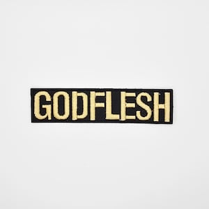 May include: A rectangular black patch with the word "GODFLESH" embroidered in gold thread. The patch is centered on a white background, showcasing the contrasting colors and bold text.