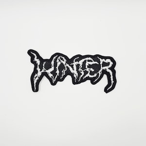 May include: A black and white embroidered patch featuring the word "WINTER" in a gothic font. The patch has a black background with white stitching for the text and border. It is designed for application on clothing or accessories.