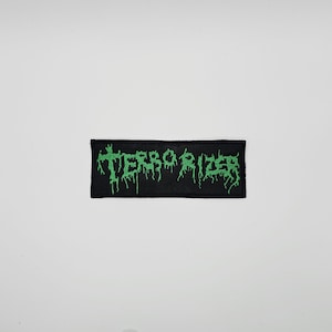 May include: Black rectangular patch with the word "TERRORIZER" embroidered in dripping green letters. The patch is likely designed to be sewn or ironed onto clothing or accessories.