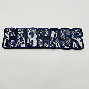 以下が含まれることがあります： 太字のブロック体で「CARCASS」という文字が刺繍された長方形のワッペン。文字は黒と白のマーブル模様または有機的なパターンで塗りつぶされており、黒い背景に対して鮮やかな青い糸の縁取りが施されています。