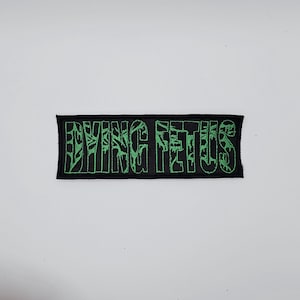 Puede incluir: Un parche rectangular negro con las palabras "DYING FETUS" bordadas en verde. El texto tiene un aspecto estilizado. Este parche es probablemente para ropa o accesorios.