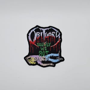 Könnte beinhalten: Schwarzer, gestickter Aufnäher mit dem Wort "Obituary" in einer stilisierten Schriftart mit roten Tropfenakzenten. Darunter steht der Text "Slowly We Rot" über einer Grafik einer verfallenden Figur. Der Aufnäher hat einen schwarzen Rand.