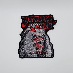 May include: Embroidered patch featuring a detailed, red-faced monster with long gray hair and an open mouth. The patch has the words "MORBID SAINT" in red, stylized lettering. The patch is black around the edges.